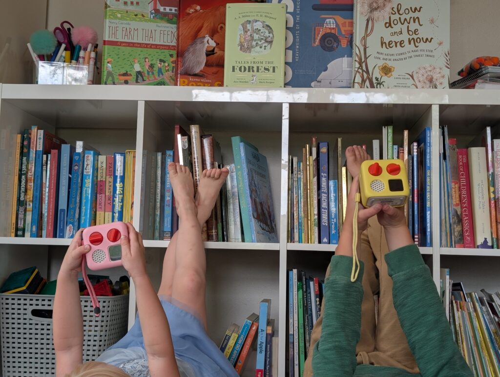 two kids holding up yoto minis, one pink and one yellow in front of a shelf of picture books